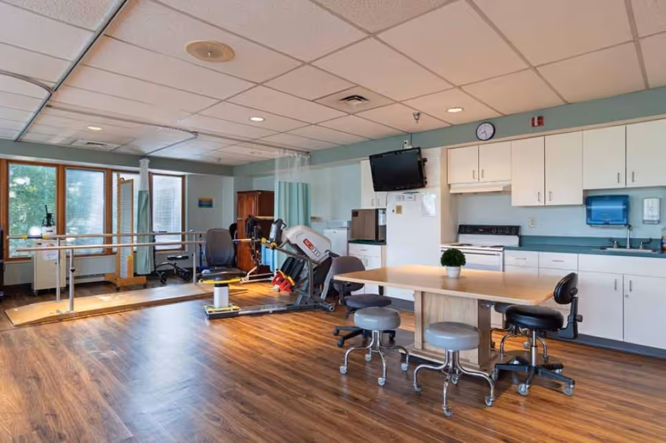A spacious room with wood flooring featuring a kitchen area with white cabinets, a stove, a sink, and a refrigerator. In the center is a wooden table surrounded by rolling stools and chairs. To the left, there is exercise equipment including parallel bars and a seated exercise machine, with large windows letting in natural light.