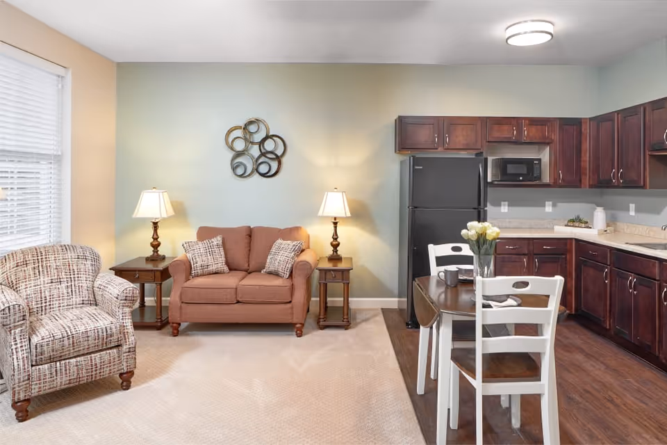 A cozy living area with a patterned armchair and a brown loveseat with two pillows, flanked by two wooden side tables each with a lamp. Adjacent to the living area is a kitchen with dark wooden cabinets, a black refrigerator, a microwave, and a small dining table with two white chairs and a vase of flowers on top.