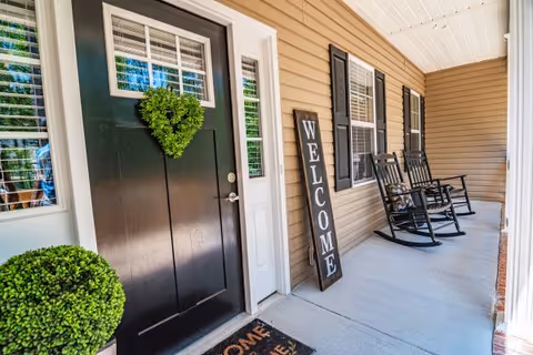 Front porch with a black entry door decorated with a heart-shaped wreath, a leaning "WELCOME" sign and two rocking chairs.