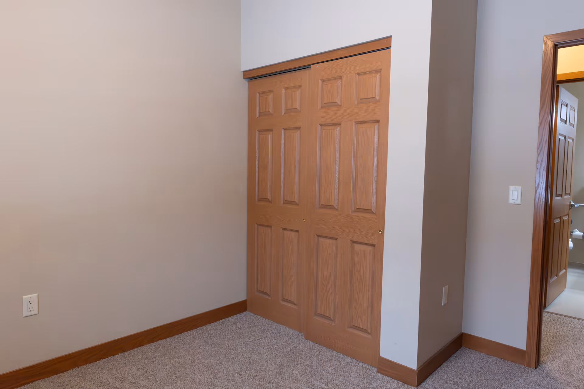 Empty room corner with beige walls, carpeted floor, and wooden trim. There is a closed wooden double closet door and an open doorway leading to another room with a visible toilet.