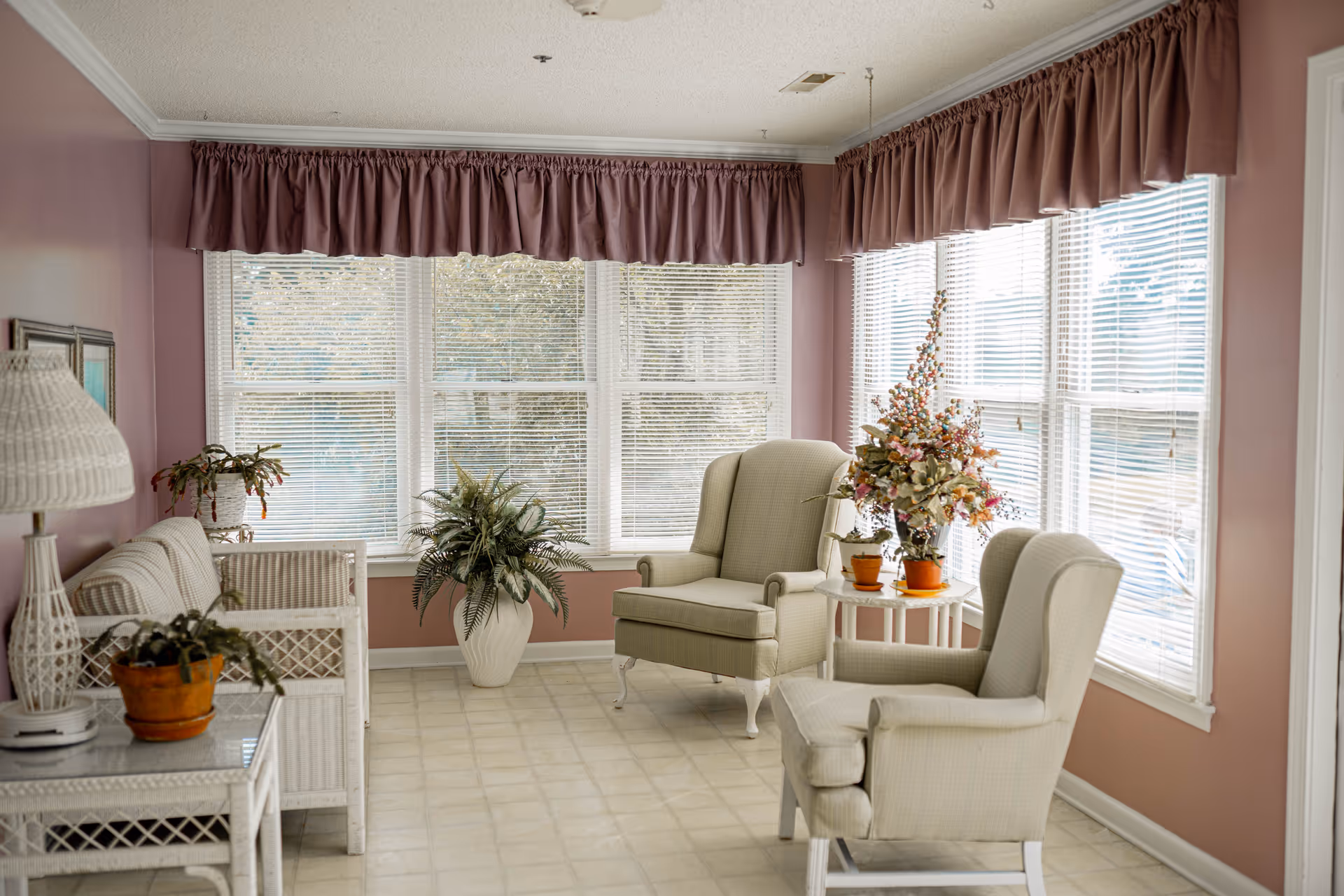 A bright sitting area with large windows covered by white blinds and mauve valances. The room features two beige wingback chairs, a white wicker loveseat, a white wicker side table with a lamp and potted plant, and a small round white table with floral arrangements. The walls are painted mauve and the floor has light-colored tiles.