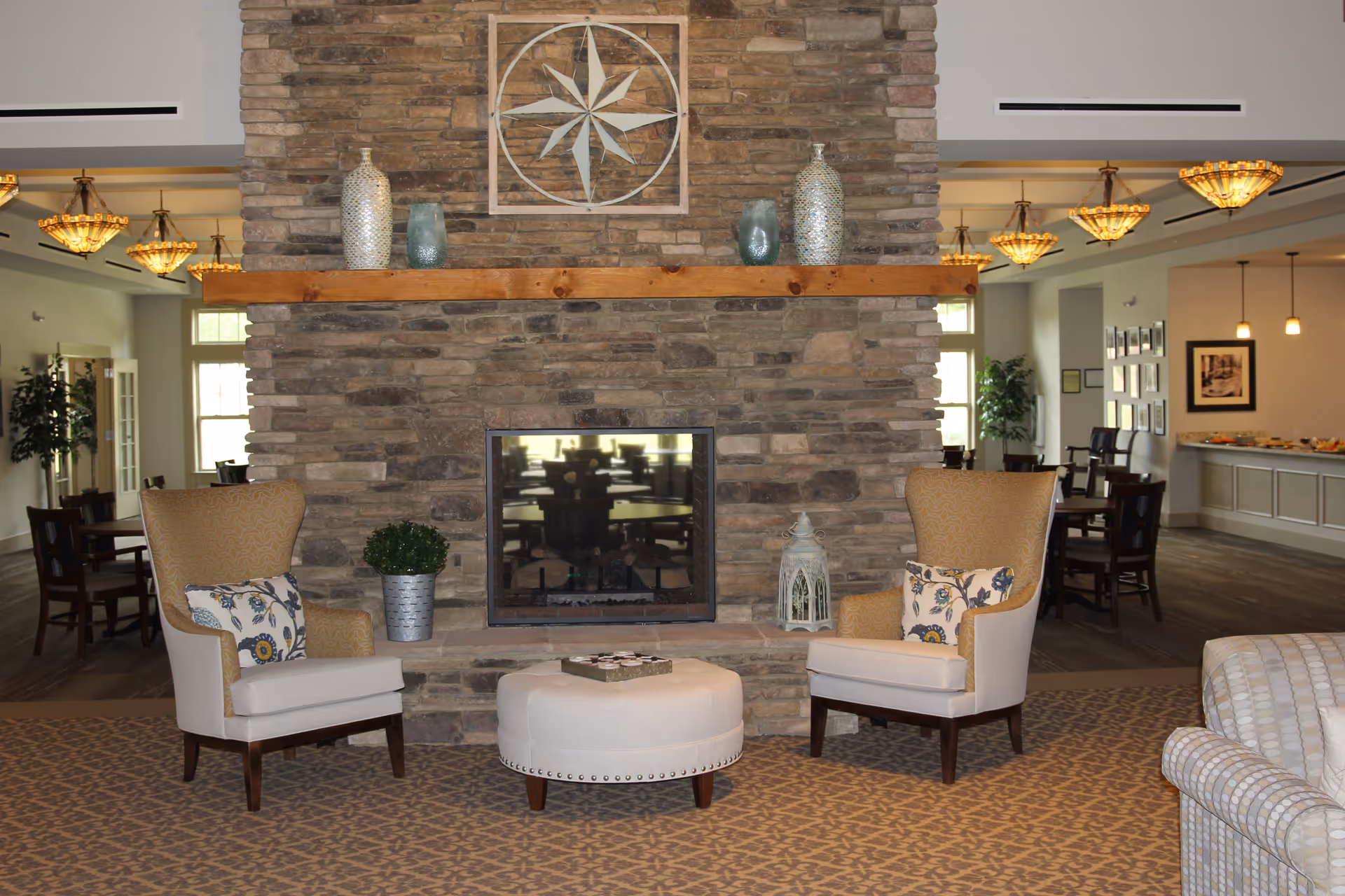 A cozy seating area in a senior living facility with two upholstered armchairs and a round ottoman in front of a large stone fireplace. Decorative vases and a lantern are placed on the wooden mantel. In the background, there are dining tables and chairs, pendant lights, and framed pictures on the walls.