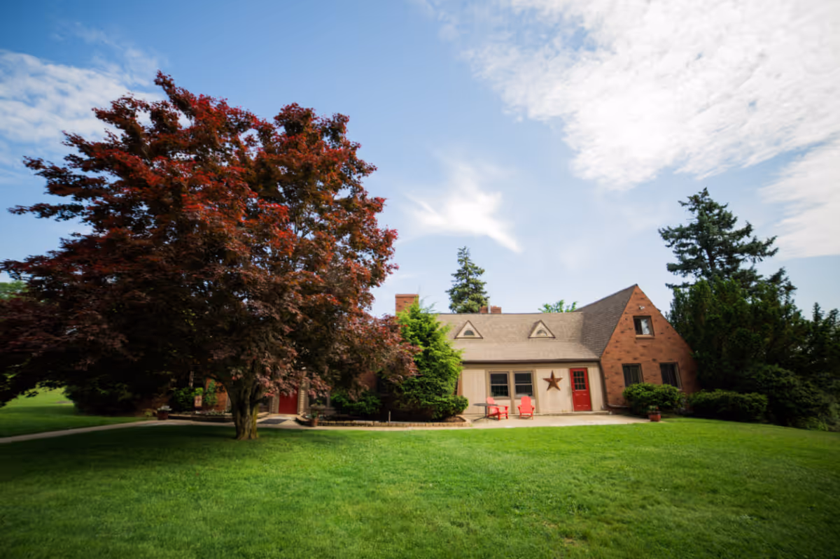 Front view of a single-story brick and siding house with a large red-leafed tree, manicured green lawn, and two red chairs on the porch under a partly cloudy sky.