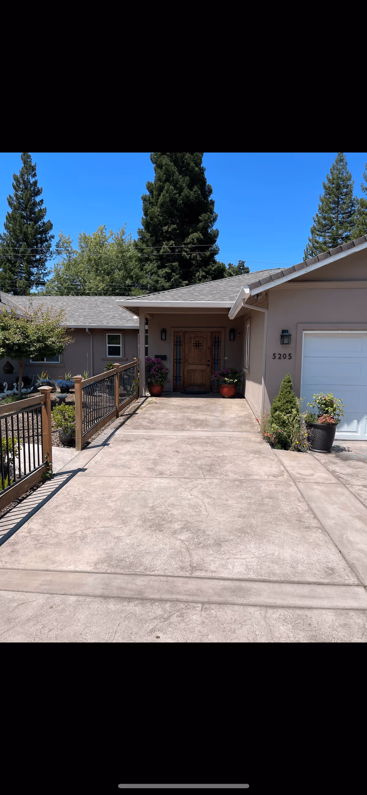 Front exterior view of a single-story house with a concrete driveway leading to a wooden front door. The house has beige walls, a gray shingled roof, and a white garage door on the right. There are potted plants with flowers on either side of the front door and along the driveway. Tall green trees and a clear blue sky are visible in the background.