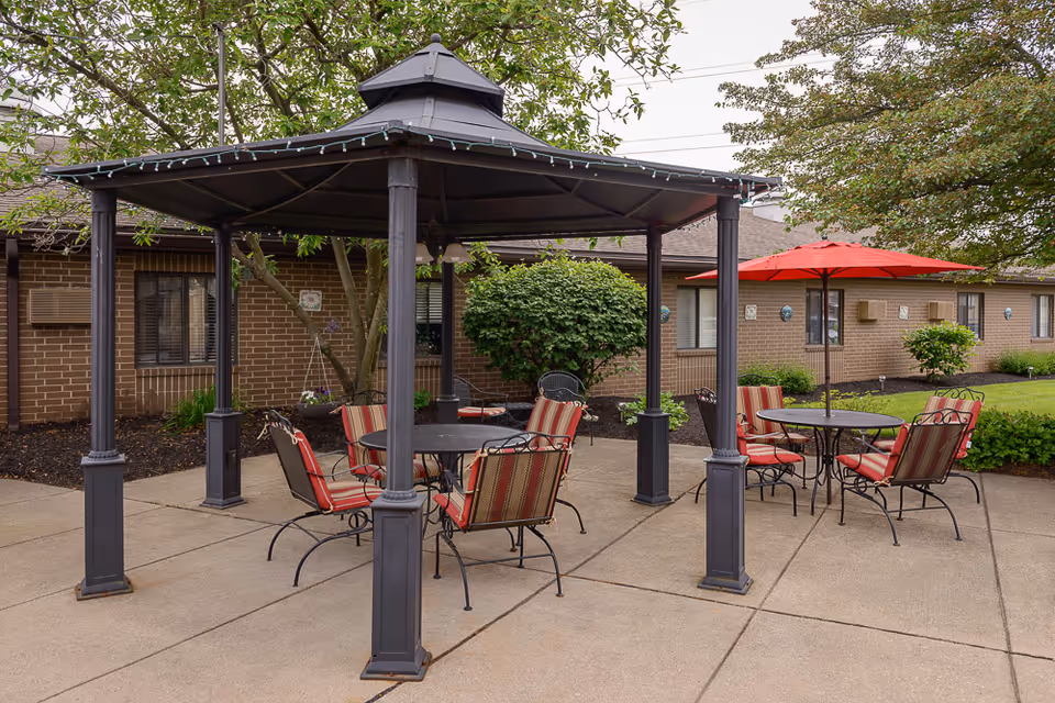 A paved outdoor patio with a gazebo, round tables, and red‑cushioned chairs in front of a brick building.