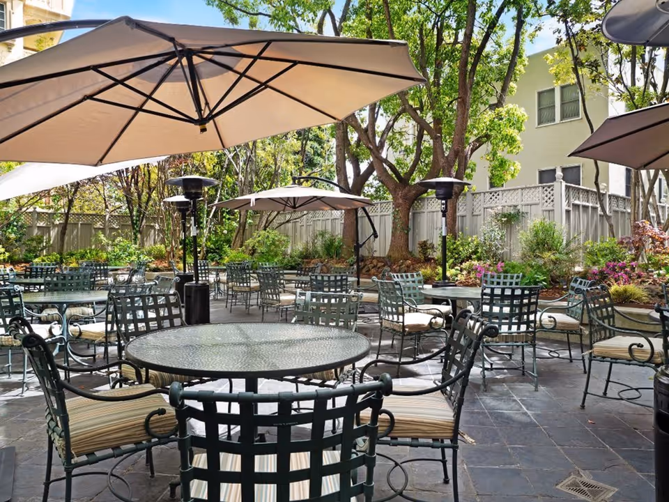 Outdoor patio area with multiple round glass tables and metal chairs with cushions, shaded by large beige umbrellas. The space is surrounded by trees, plants, and a wooden fence, with a building visible in the background.