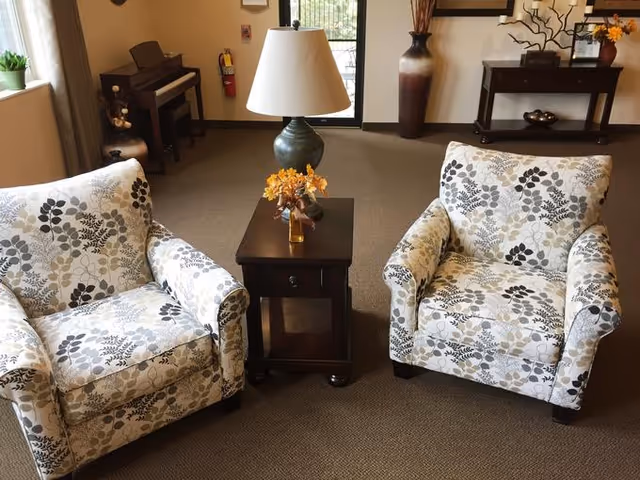 A cozy sitting area with two floral-patterned armchairs facing each other with a small wooden side table in between. The table holds a lamp with a beige shade and a small vase with orange flowers. In the background, there is a piano, a fire extinguisher mounted on the wall, a large decorative vase, and a console table with decorative items. The room has beige carpeting and a window letting in natural light.