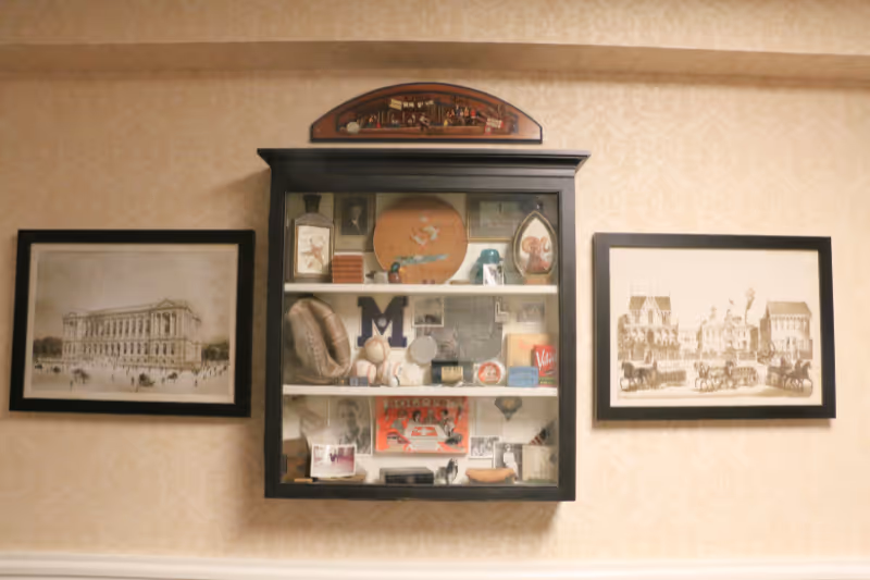 A wall display featuring a black-framed glass cabinet filled with various vintage memorabilia and collectibles, flanked by two framed sepia-toned prints of historic buildings on either side. The wall has a beige patterned wallpaper background.