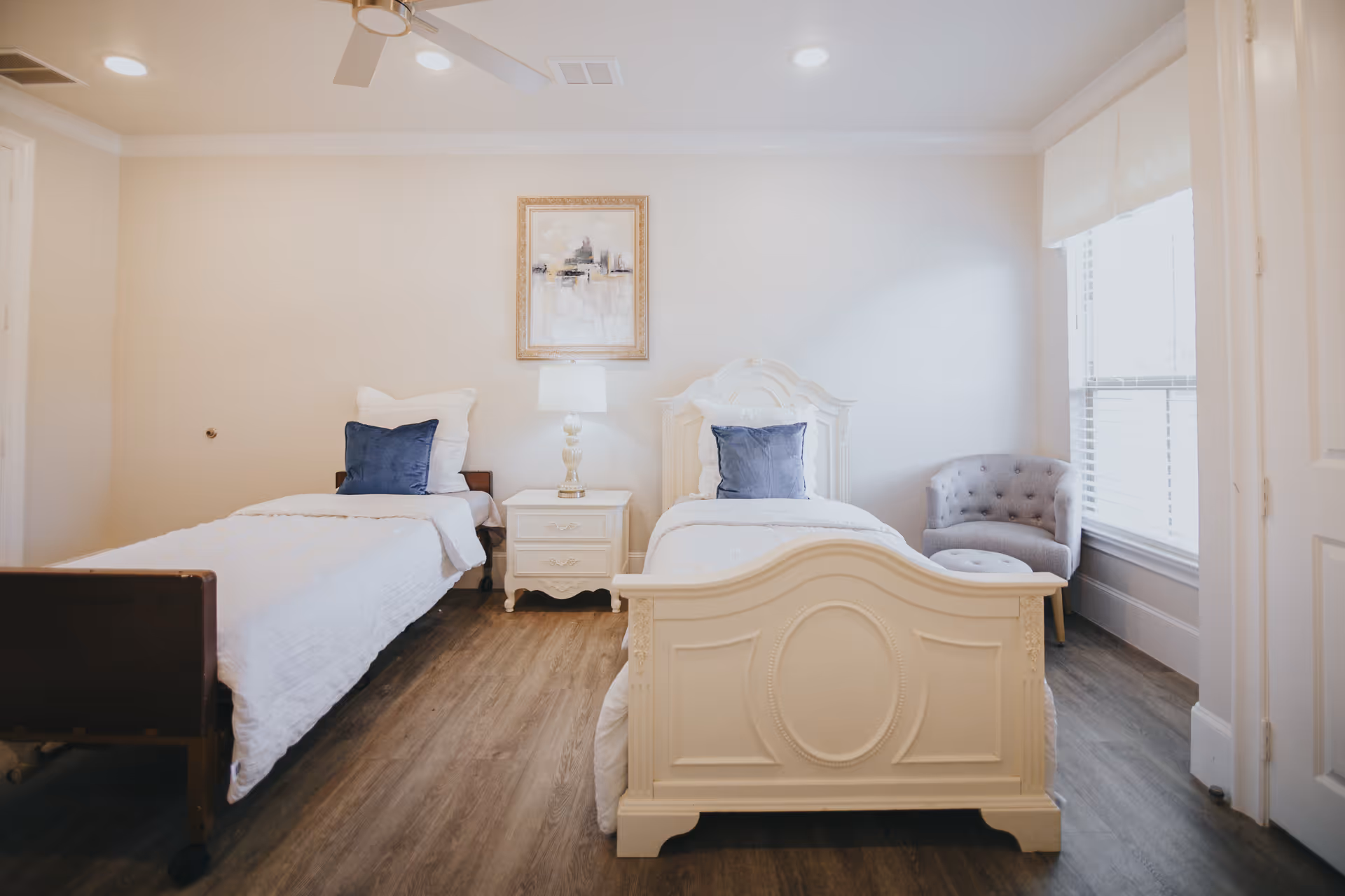 A bright bedroom with two single beds, each with white bedding and a blue pillow. Between the beds is a white nightstand with a lamp and a framed picture above it. To the right, there is a window with blinds and a gray upholstered chair with a matching ottoman. The floor is wood, and the ceiling has a white fan.