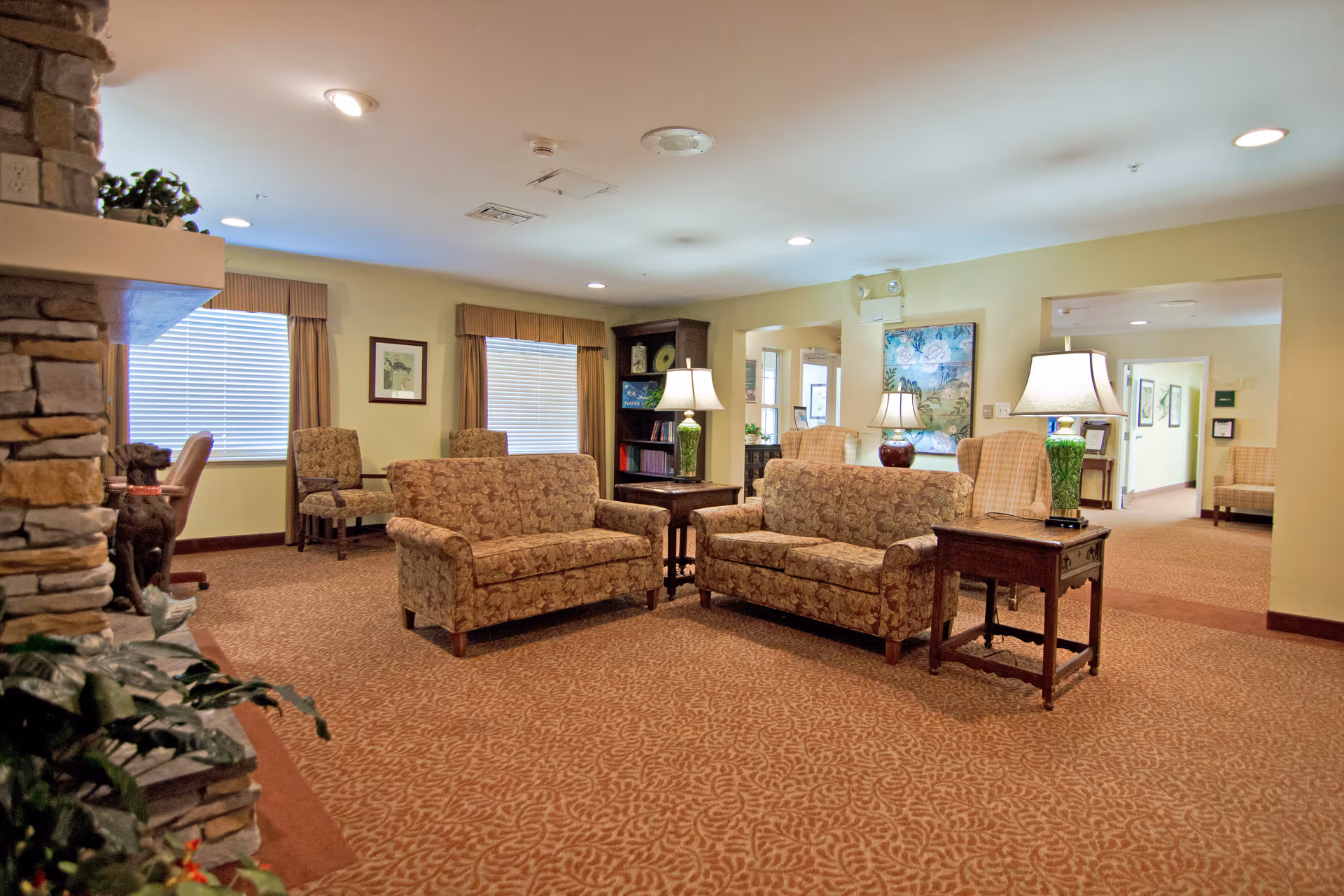 A comfortable senior living lounge with patterned sofas, side tables and lamps, carpeting and a stone fireplace.