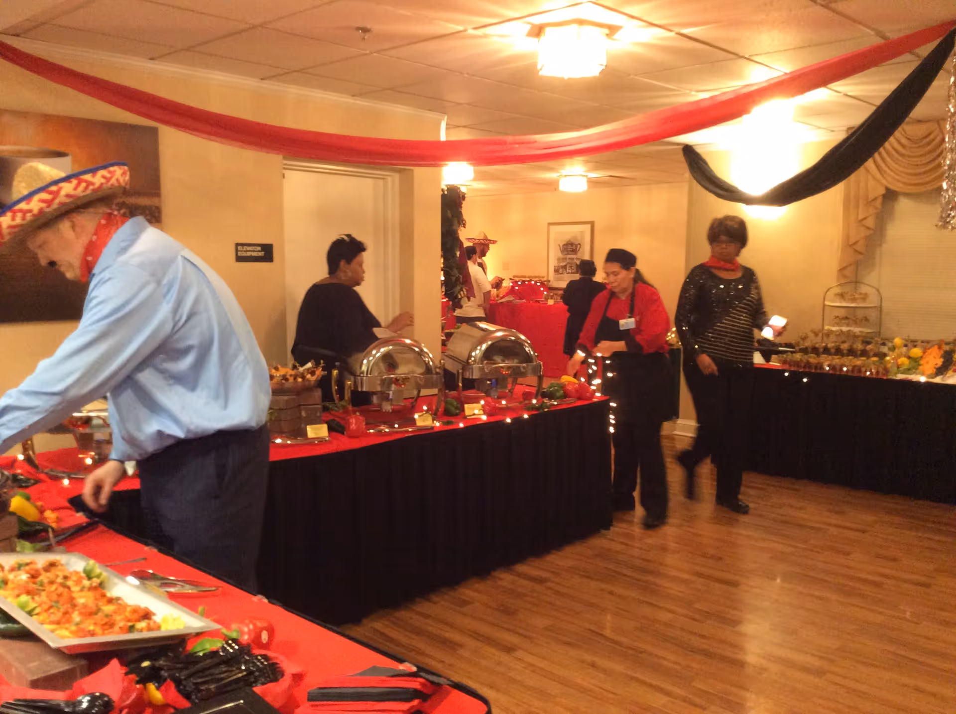 A buffet setup in an indoor room decorated with red and black drapes hanging from the ceiling. Several people are serving or preparing food on tables covered with red and black tablecloths. The floor is wooden, and the lighting is warm with ceiling lights.