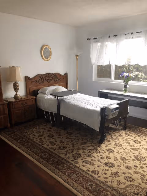 Furnished bedroom with a single adjustable bed, ornate wooden headboard, bedside table, lamp and a window with flowers.