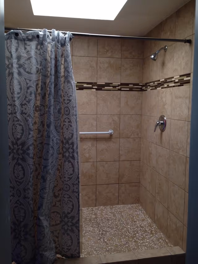 A tiled shower area with a patterned shower curtain partially drawn to the left. The shower walls are covered with beige tiles and a decorative horizontal strip of smaller rectangular tiles. There is a silver showerhead and a single handle control on the right wall, and a white grab bar mounted on the back wall. The shower floor has small pebble-like tiles, and a skylight above provides natural light.