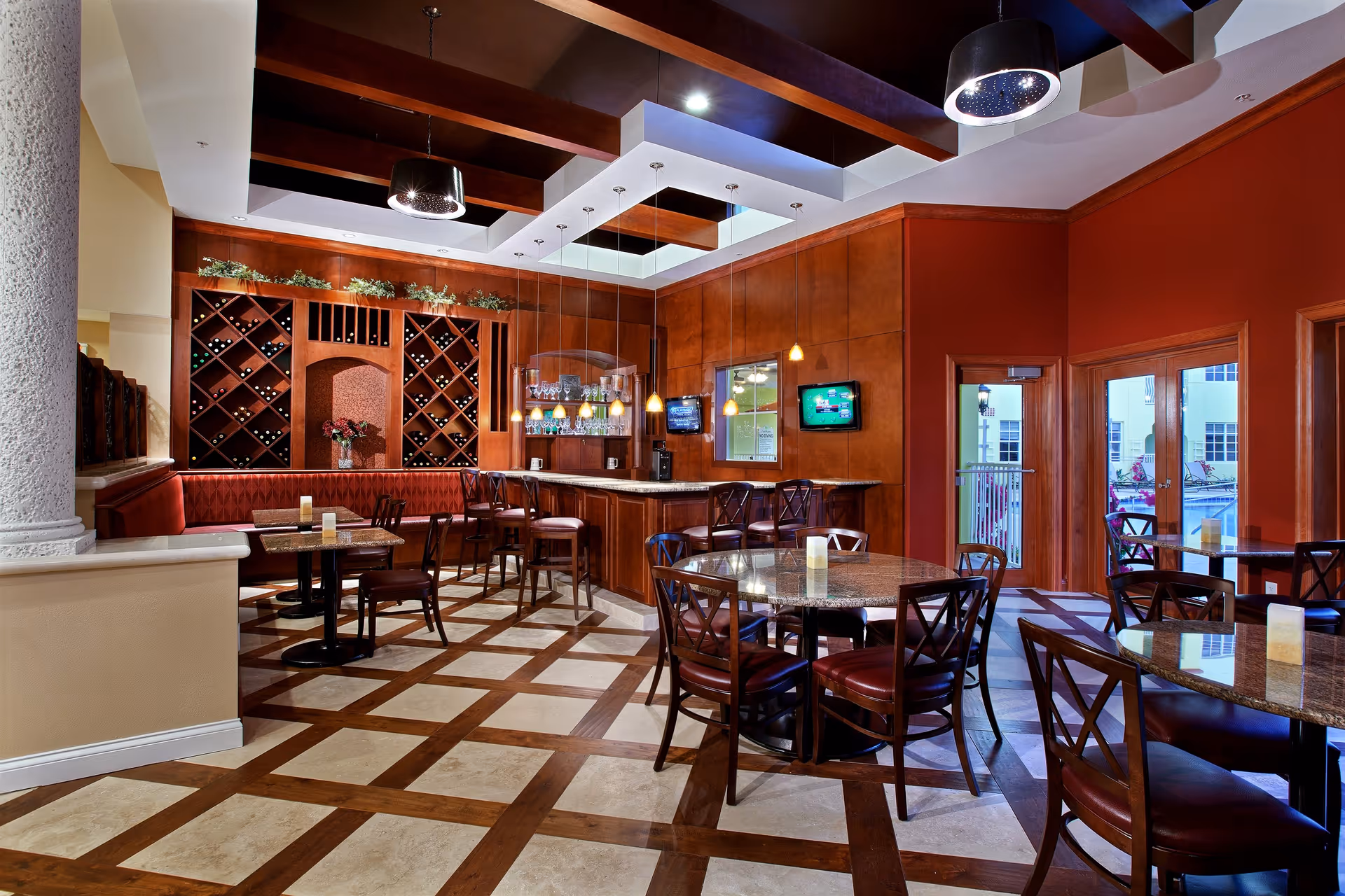 Interior view of a dining area with multiple round tables and chairs, a bar with high stools, wine racks on the wall, pendant lights hanging from the ceiling, and large windows and doors leading outside to a pool area.