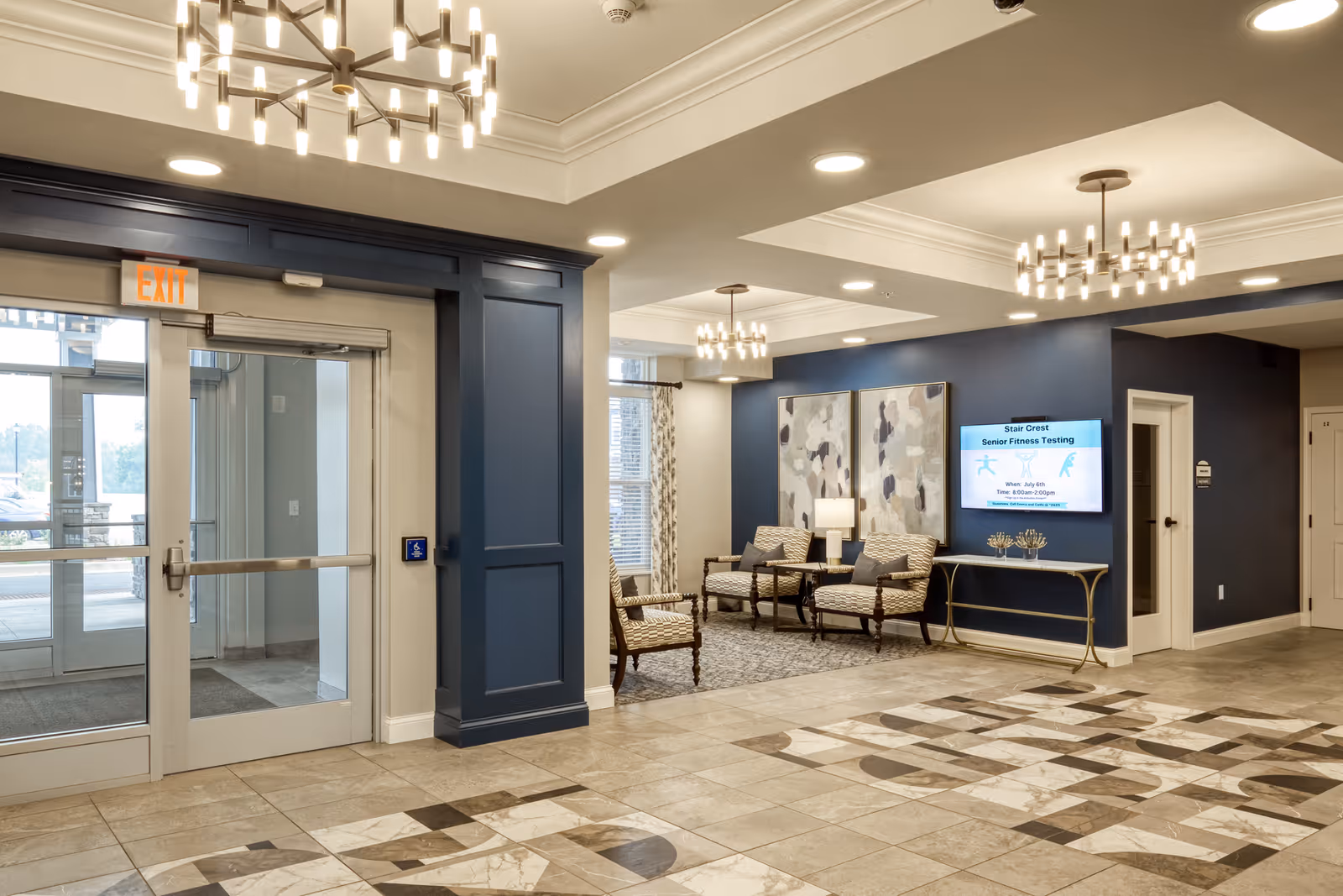 Interior view of a senior living facility lobby at Stair Crest featuring a tiled floor with geometric patterns, a glass exit door, blue accent walls, modern chandeliers, and a seating area with patterned armchairs and a small table. A digital screen on the wall displays information about senior fitness testing.