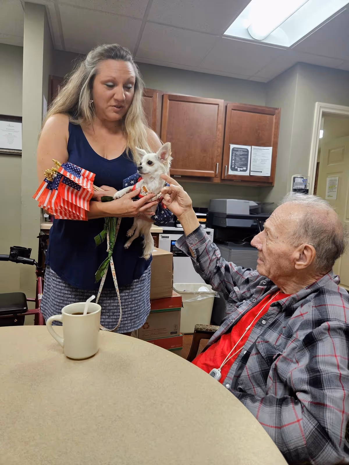 An elderly man sitting at a table reaches out to pet a small white dog held by a woman standing next to him. The woman is holding several small American flags and the setting appears to be a room with wooden cabinets and office equipment in the background.
