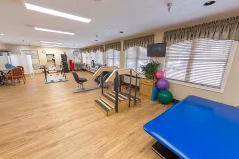 Bright rehabilitation gym with therapy stairs, treatment tables, exercise balls and large windows along one wall.