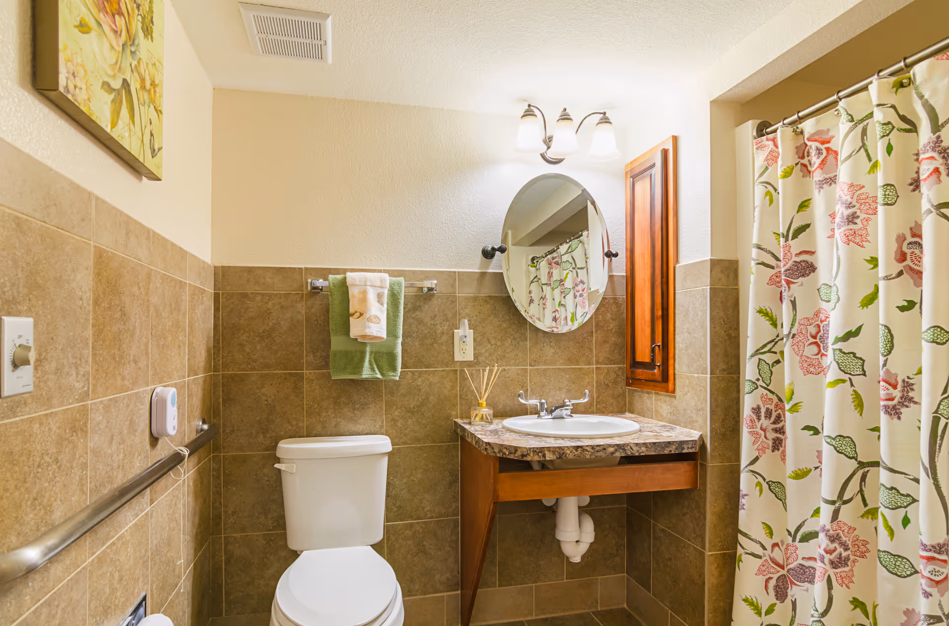 A clean and well-lit bathroom featuring a white toilet, a sink with a marble countertop, an oval mirror above the sink, a wooden cabinet mounted on the wall, and a shower with a floral-patterned curtain. The walls are tiled halfway up with brown tiles and painted cream above. A towel rack with green and white towels is mounted above the toilet, and a floral painting hangs on the wall.
