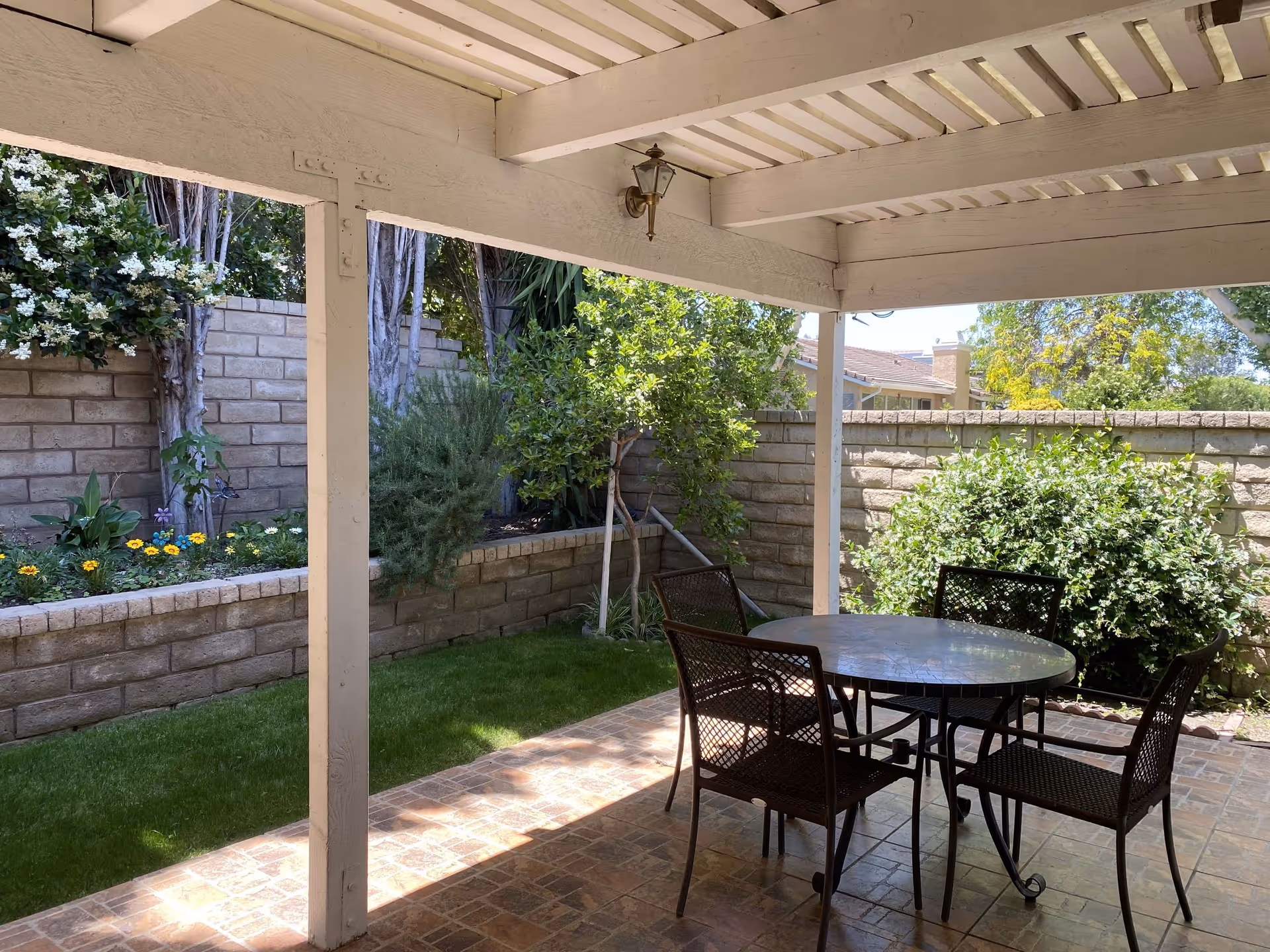 Covered patio with a round table and four chairs next to a small fenced garden and raised flower beds.