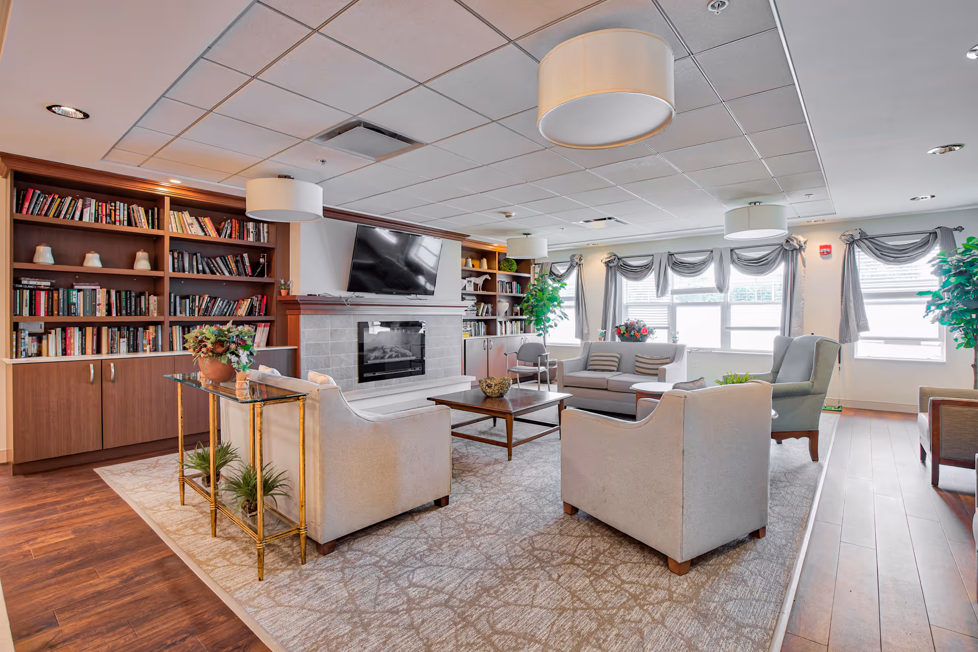 A bright and spacious living room area in a senior living facility featuring comfortable beige armchairs and a sofa arranged around a wooden coffee table on a patterned rug. The room has a fireplace with a mounted flat-screen TV above it, built-in bookshelves filled with books and decorative items, large windows with gray curtains allowing natural light, and modern ceiling lights. There are also green plants adding a touch of nature to the space.