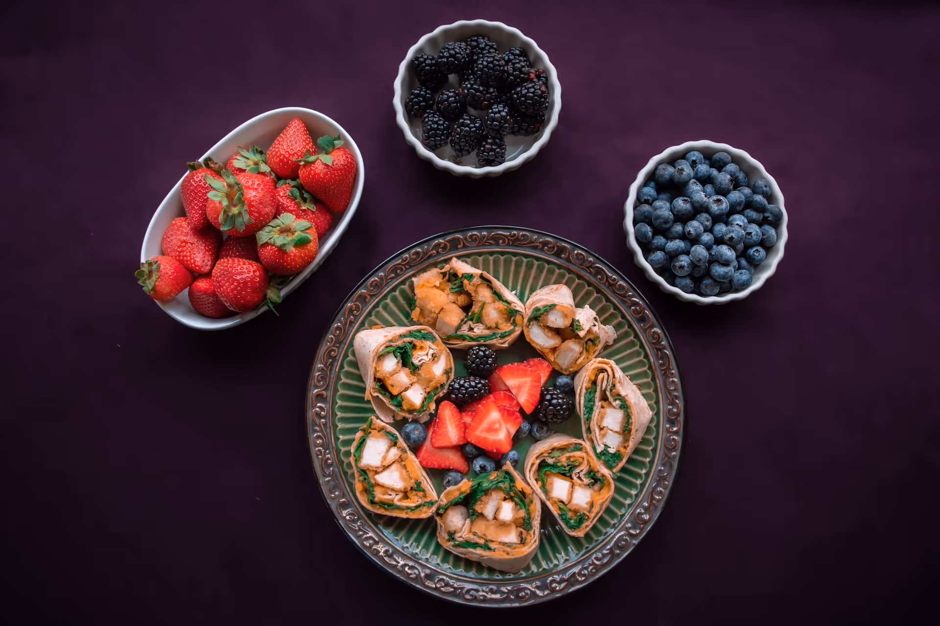 A green decorative plate with several pieces of chicken wrap sandwiches arranged in a circle, garnished with strawberries, blackberries, and blueberries in the center. Surrounding the plate are three small bowls containing fresh strawberries, blackberries, and blueberries on a dark purple tablecloth.