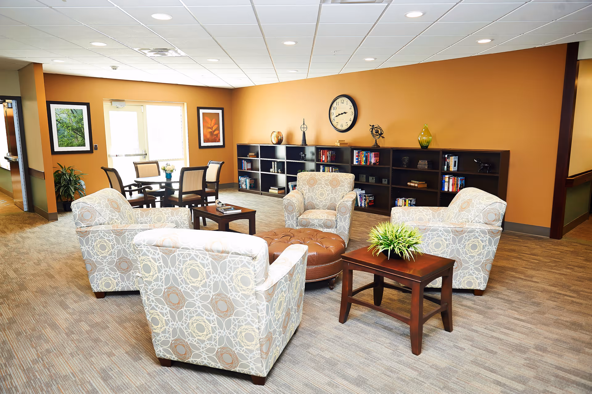 A cozy senior living common area with four patterned armchairs arranged around a round brown ottoman. There are two wooden side tables, one with a green plant. In the background, there is a long black bookshelf with books and decorative items, a wall clock, and two framed pictures on an orange wall. A small dining table with four chairs is near a glass door letting in natural light.