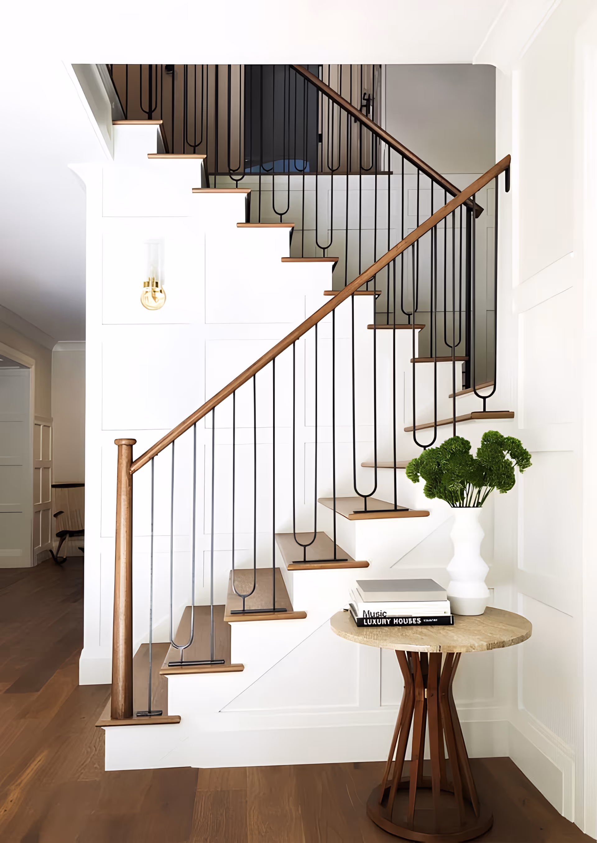A modern interior staircase with wooden steps and black metal railings. Next to the staircase is a round wooden table with a white vase holding green leafy plants and a stack of books. The walls are white, and the floor is wooden.