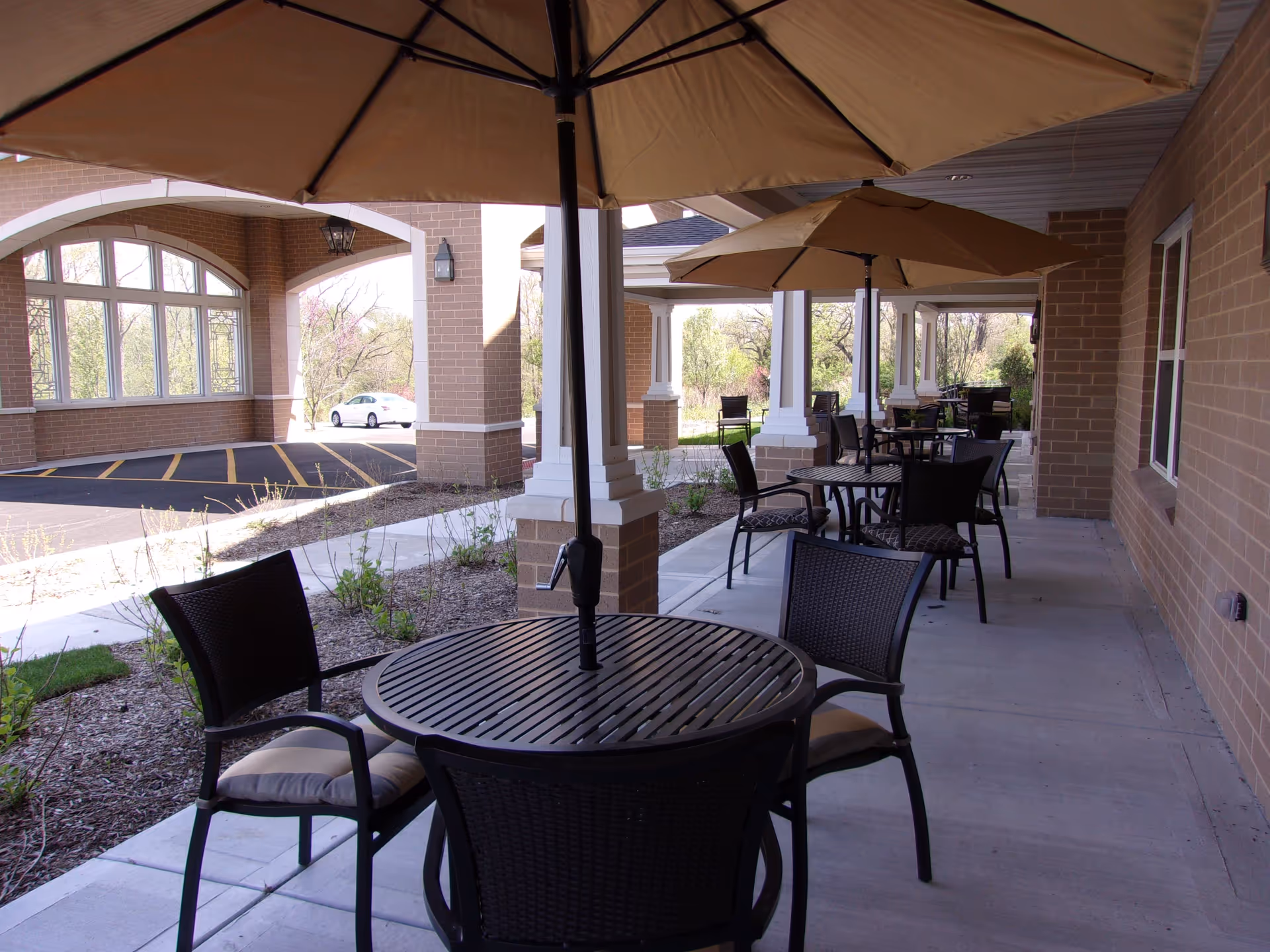 Outdoor patio area with round tables and chairs under large beige umbrellas, adjacent to a brick building with columns and windows. There is a parking area visible in the background with a white car parked.