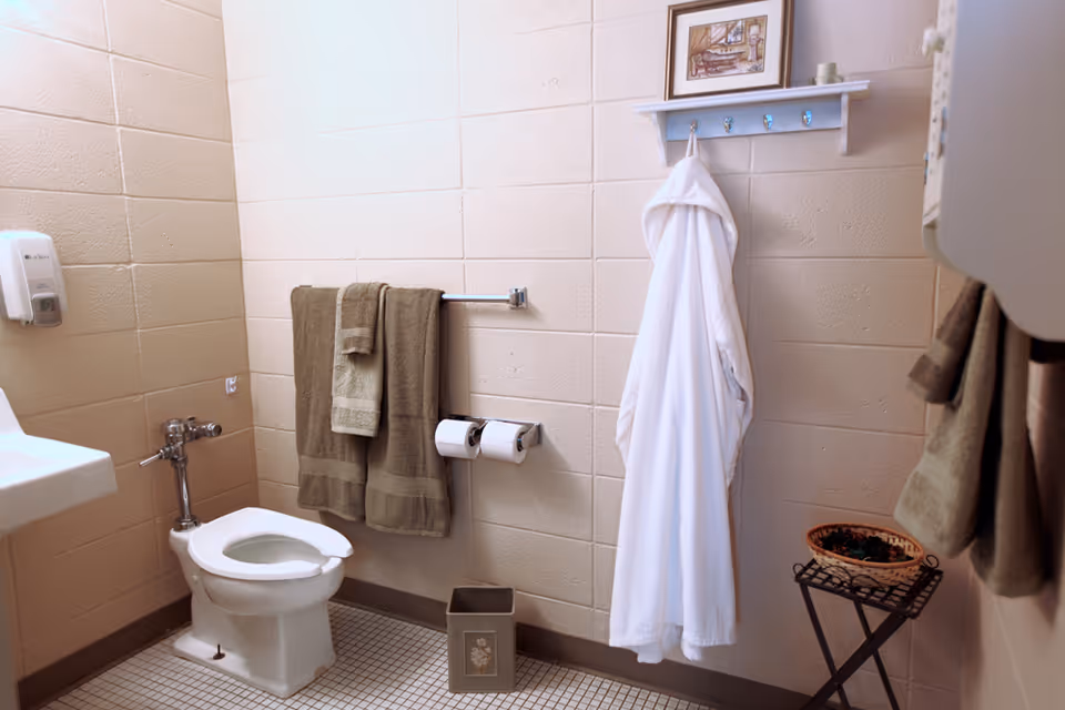 A clean bathroom with a white toilet, a white sink, beige tiled walls, and beige floor tiles. Two brown towels hang on a towel rack above the toilet paper holder with two rolls. A white bathrobe hangs on a wall-mounted rack with hooks, above a small decorative trash bin and a small table with a basket. A framed picture is mounted above the bathrobe.