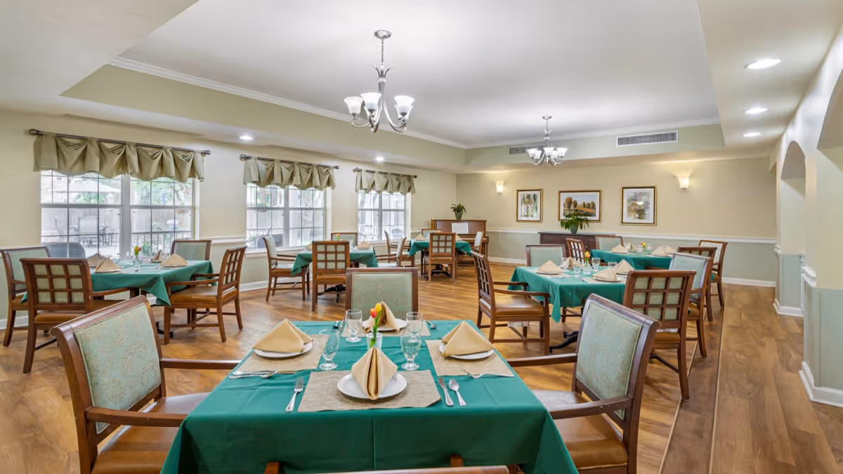 Bright dining room with several tables set with green tablecloths, place settings, wooden chairs, and chandeliers.