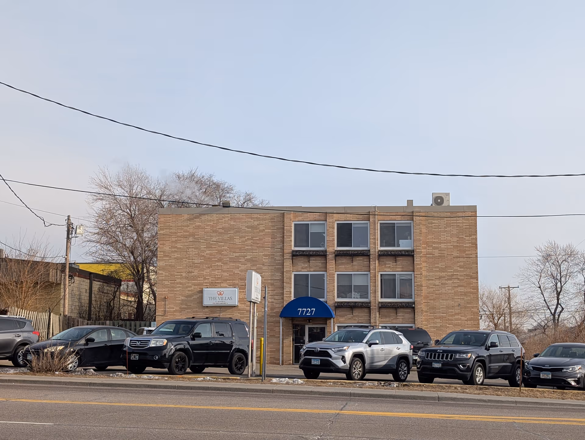 Three-story brick apartment building with a blue awning reading "7727", a sign for The Villas, and cars parked in front.