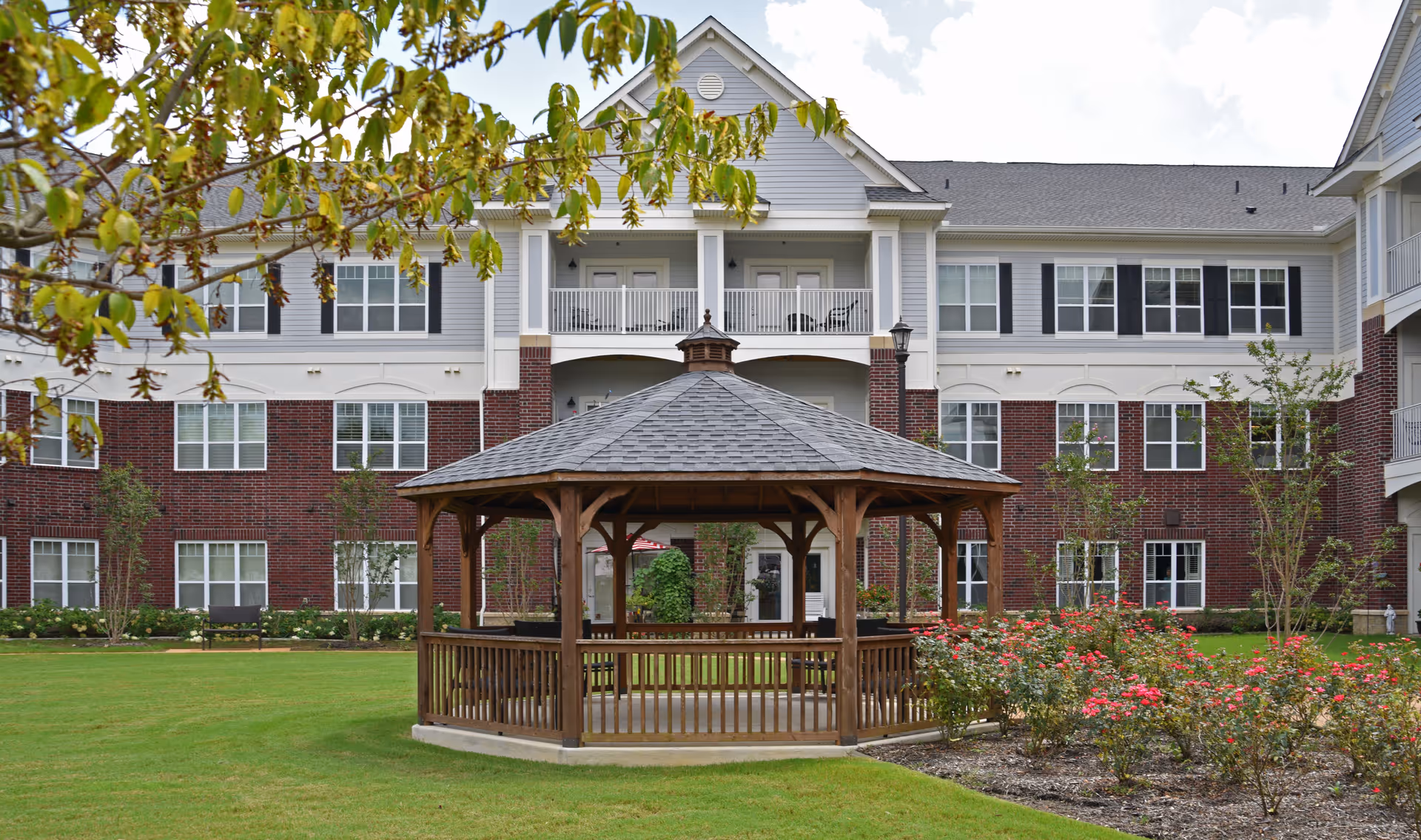 A spacious outdoor garden area at Irene Woods Assisted Living featuring a wooden gazebo in the center, surrounded by green grass, flowering bushes, and trees. The background shows a multi-story brick and siding building with balconies and windows.