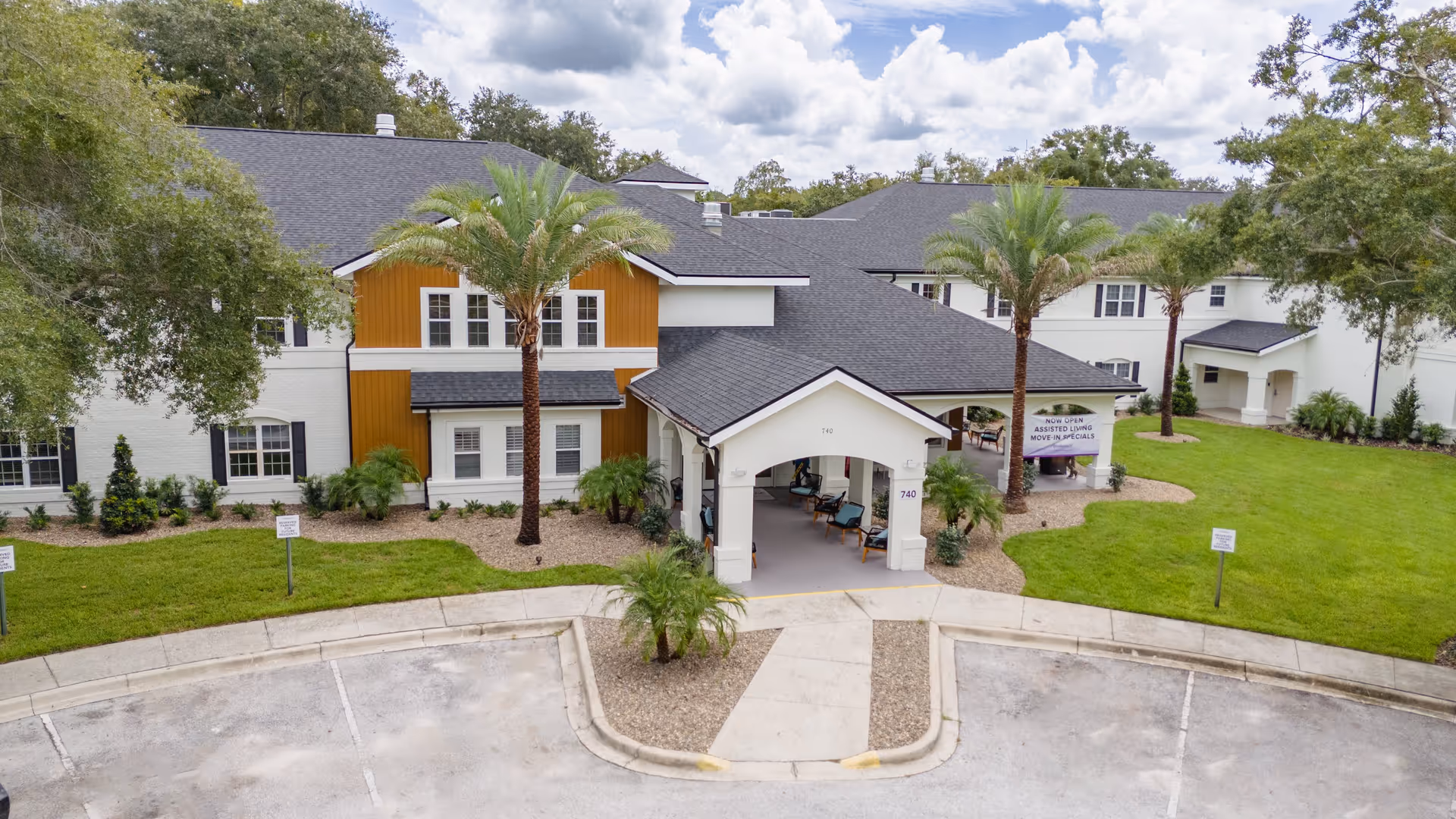 Front exterior view of Ambiance at Maitland facility showing a two-story building with white and orange siding, black roof, palm trees, green lawn, and a covered entrance with seating. A sign near the entrance reads 'Now Open Assisted Living Move-In Specials'.