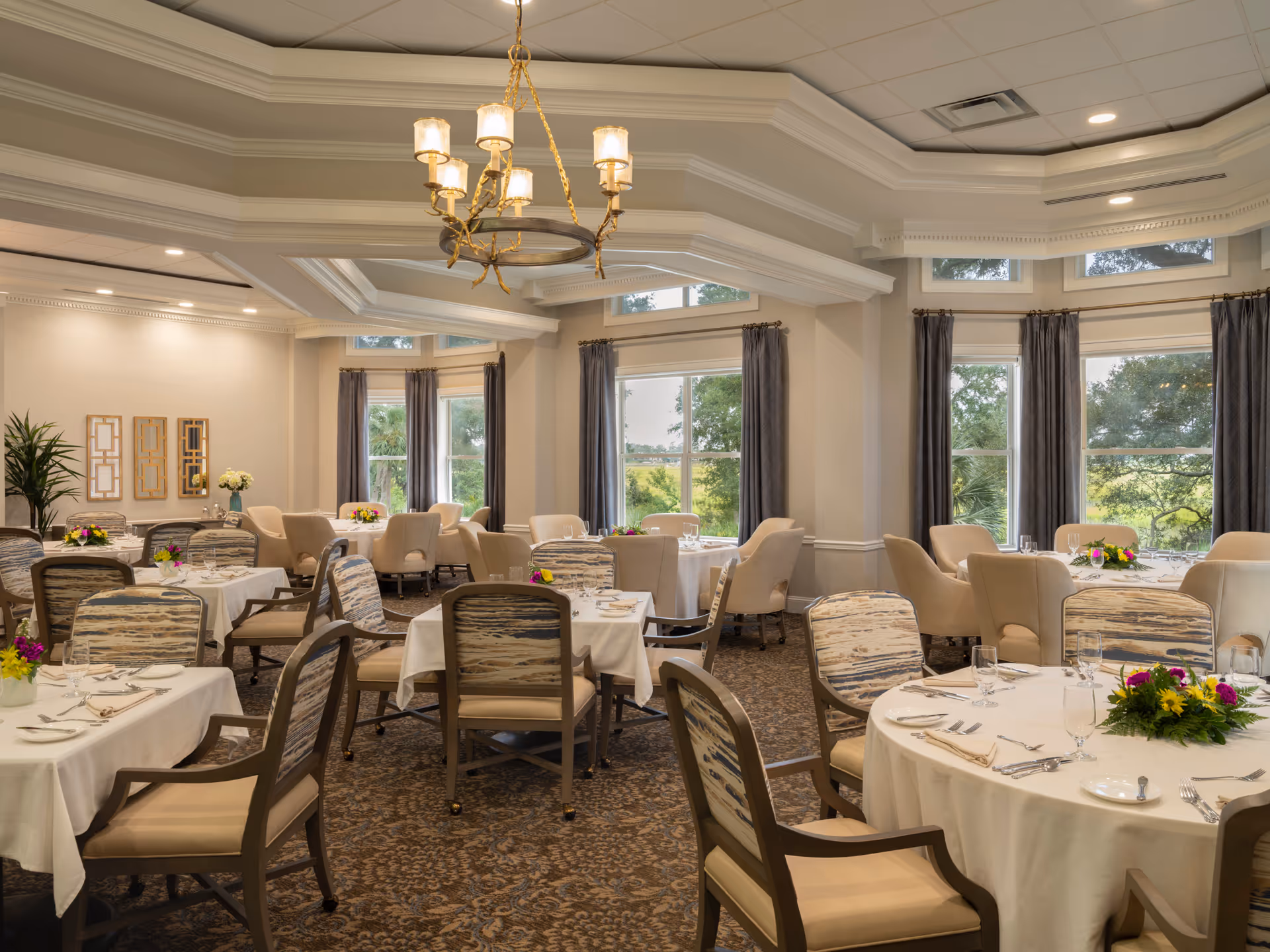 Elegant dining room with round and rectangular tables set with white linens and floral centerpieces, upholstered chairs, large windows and a chandelier.