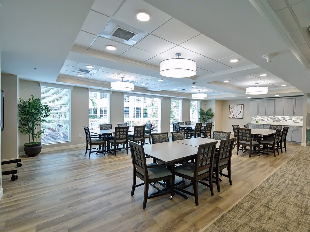 Bright communal dining area with multiple tables and chairs, large windows, pendant lighting, and a kitchenette.
