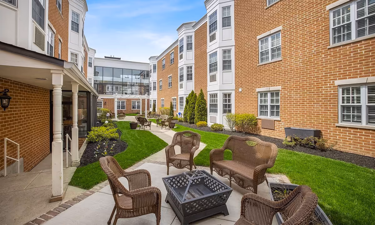 Outdoor courtyard area at Overlook Village featuring wicker chairs and a loveseat arranged around a square fire pit on a paved patio. The courtyard is surrounded by brick buildings with multiple windows and landscaped with green grass, shrubs, and small trees. A covered walkway with white columns is visible on the left side.