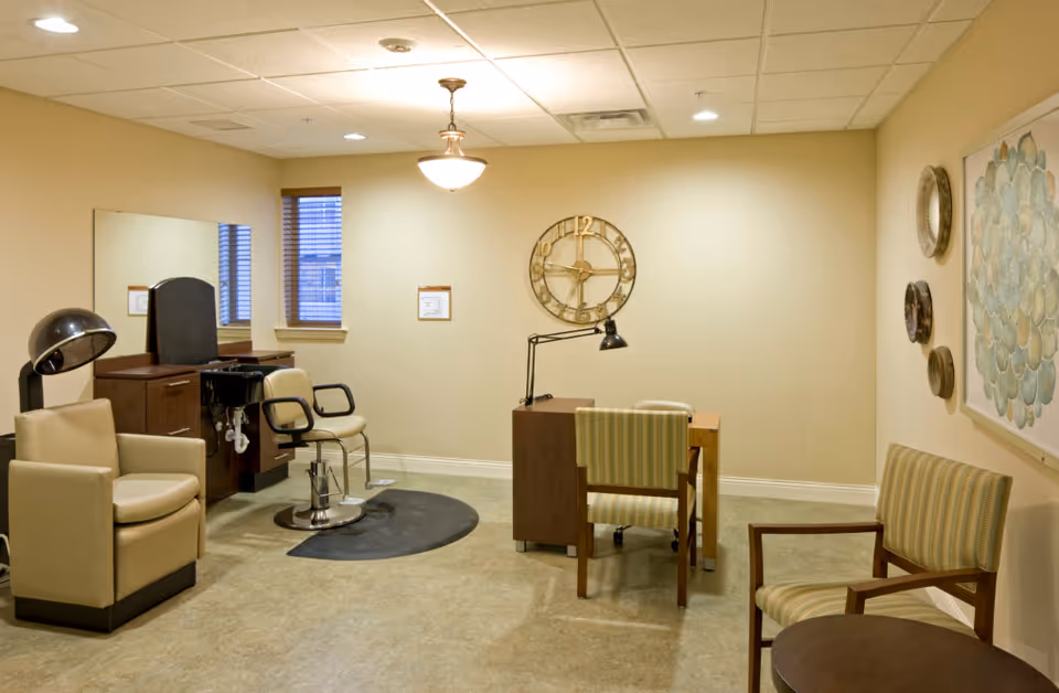 Interior of a salon room in a senior living facility featuring a hair drying chair with a hooded dryer, a styling chair in front of a mirror, a small desk with a lamp and two chairs, wall clock, and decorative wall art.
