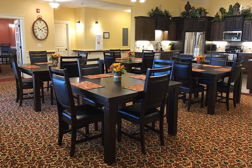 Dining room with several dark wood tables and chairs set with placemats and small floral centerpieces, with a kitchenette in the background.