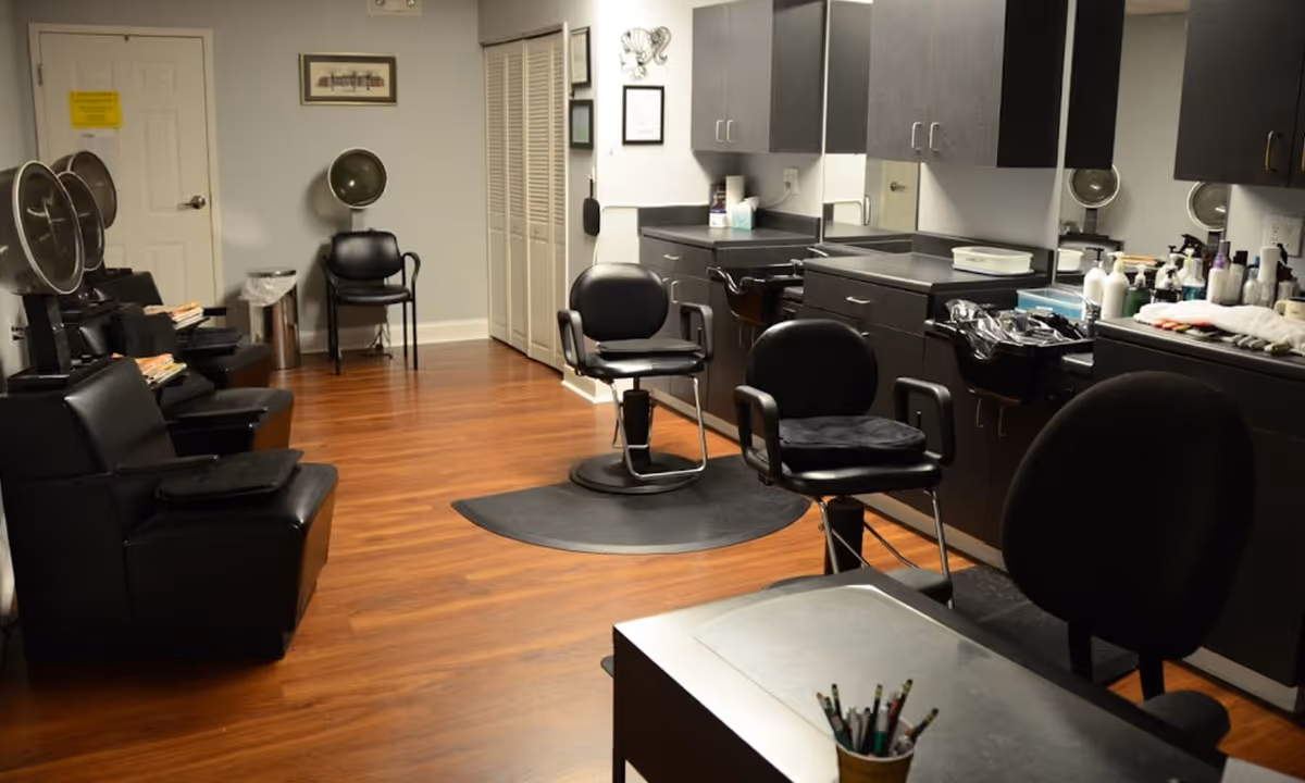 Interior view of a salon area in a senior living facility with black salon chairs, hair dryers, and cabinets along the wall. The room has wooden flooring and a small table with pencils in the foreground.