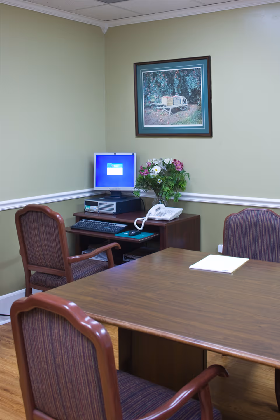 A small office or meeting room with a wooden table and three upholstered chairs. In the corner, there is a desk with a desktop computer, a telephone, and a vase with flowers. A framed picture of a wheelbarrow with apples hangs on the light green wall.