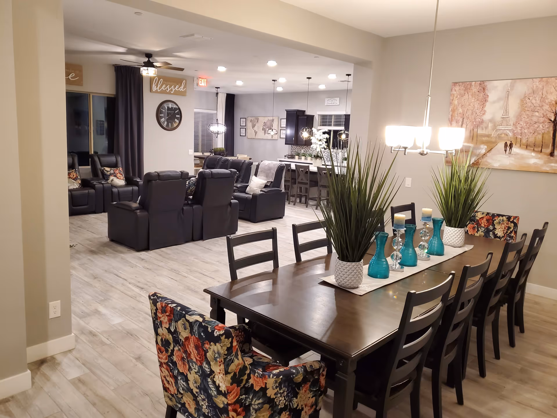 A spacious and well-lit interior of a senior living facility featuring a long wooden dining table with floral and black chairs, decorated with green plants and blue vases. In the background, there is a living area with black recliner chairs, a wall clock, and decorative signs. The kitchen area with bar stools and pendant lights is also visible.