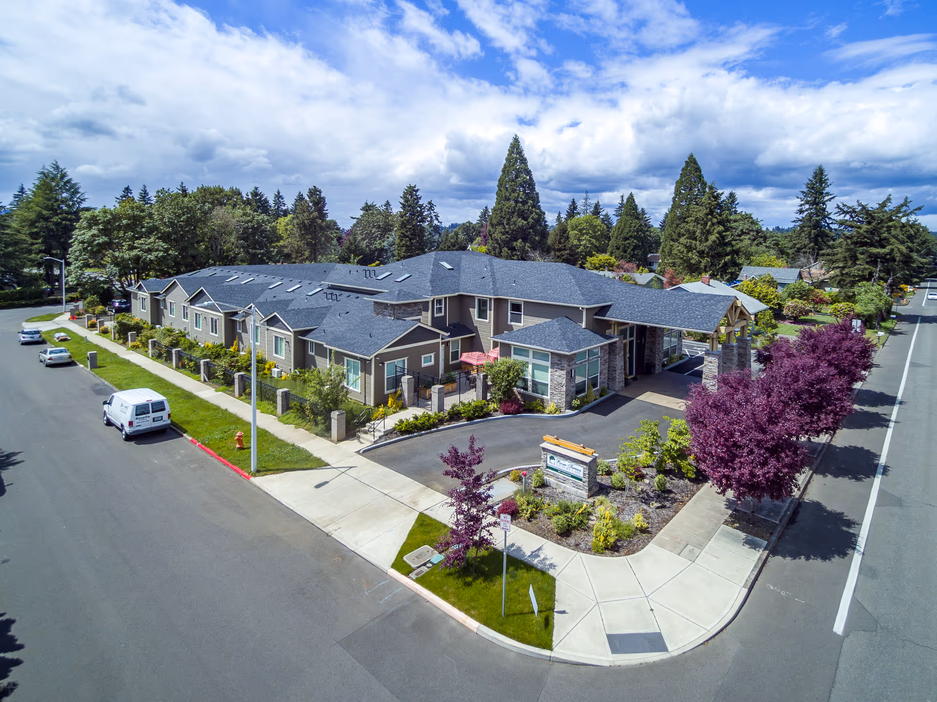 Aerial view of River Terrace Memory Care facility showing a single-story building with a dark roof, surrounded by landscaped greenery and trees. There is a driveway leading to a covered entrance, and several cars are parked along the street adjacent to the building. The sky is partly cloudy with patches of blue.