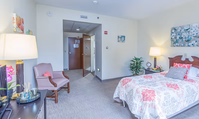 Furnished assisted-living bedroom with a bed, nightstand, armchair, lamps, and an open doorway to a hallway.
