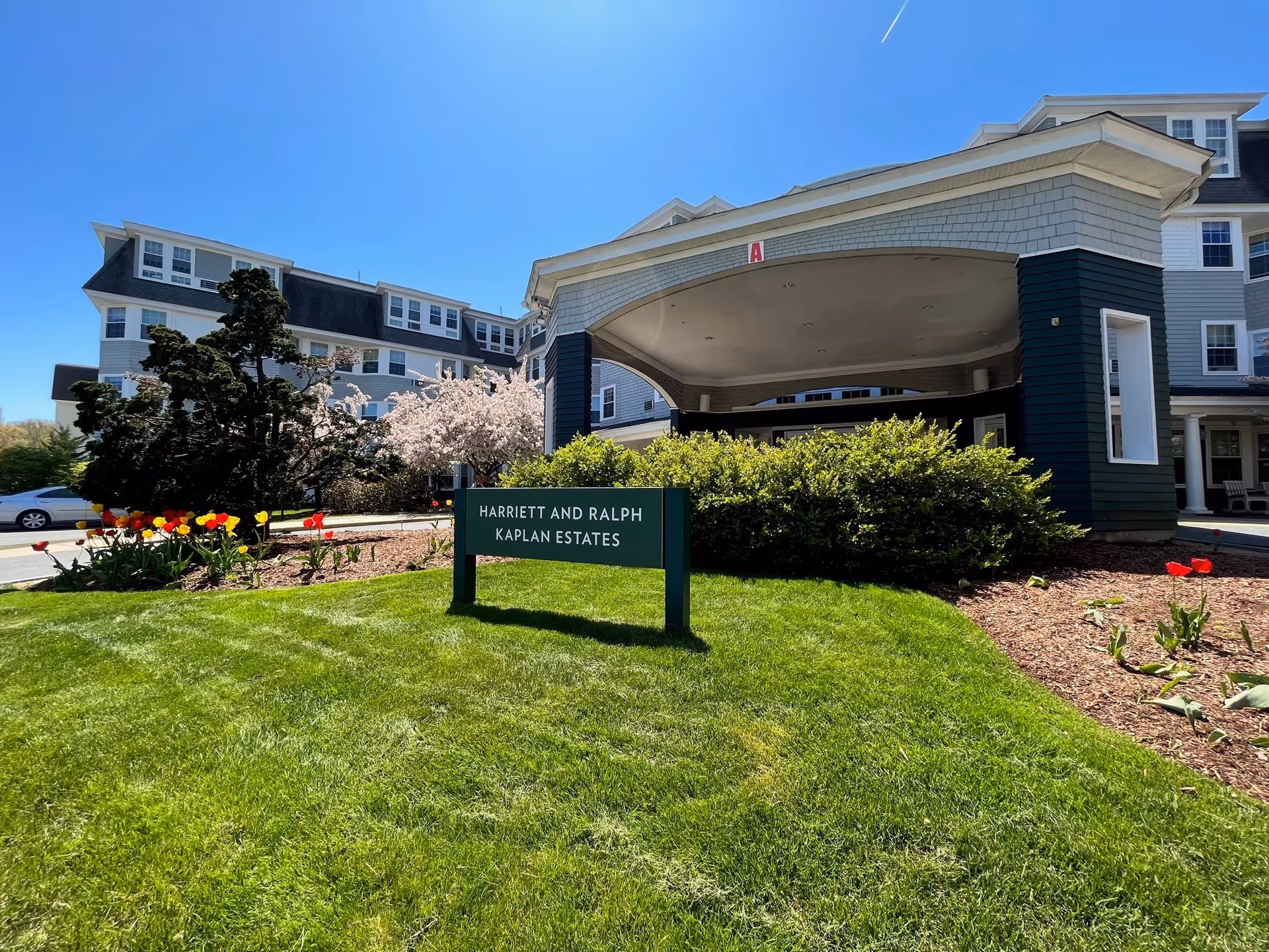 Exterior view of the Legacy at Harriett & Ralph Kaplan Estates building on a sunny day, showing a green lawn with colorful tulips and a sign that reads 'Harriett and Ralph Kaplan Estates' in front of the entrance canopy.