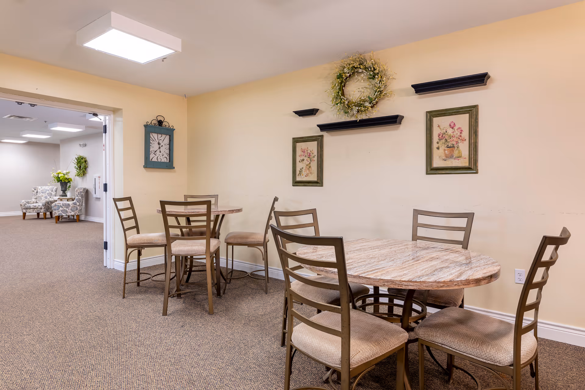 A cozy interior common area with two round tables and several chairs arranged around them. The walls are painted light yellow and decorated with two framed floral paintings, a green wreath, and three black floating shelves. In the background, there is an open doorway leading to another room with two patterned armchairs and a small table with a flower arrangement.