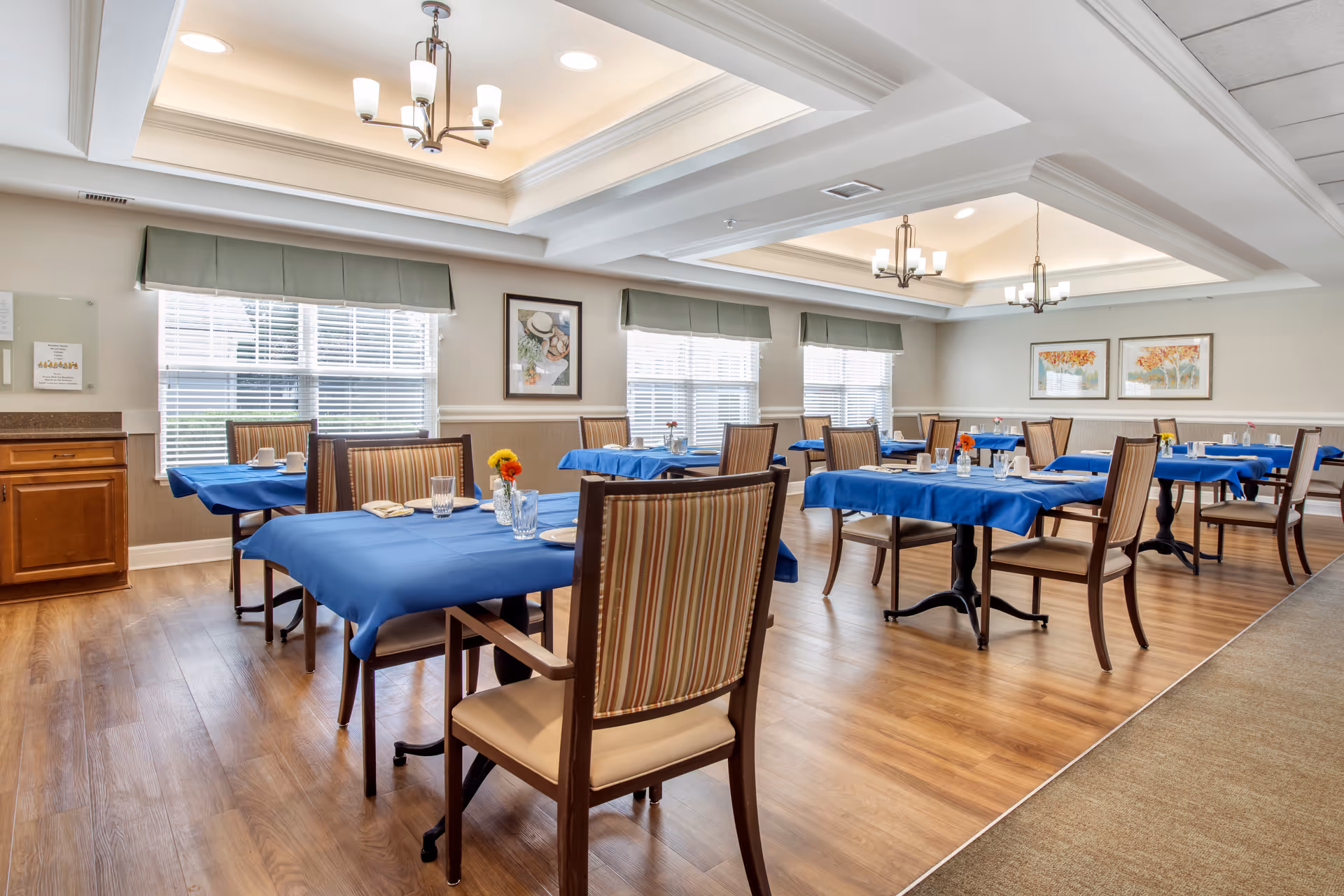 A bright dining room with wooden floors and multiple tables covered with blue tablecloths. Each table is set with plates, glasses, napkins, and small flower vases. The room has large windows with blinds and green valances, framed artwork on the walls, and ceiling lights with modern chandeliers.