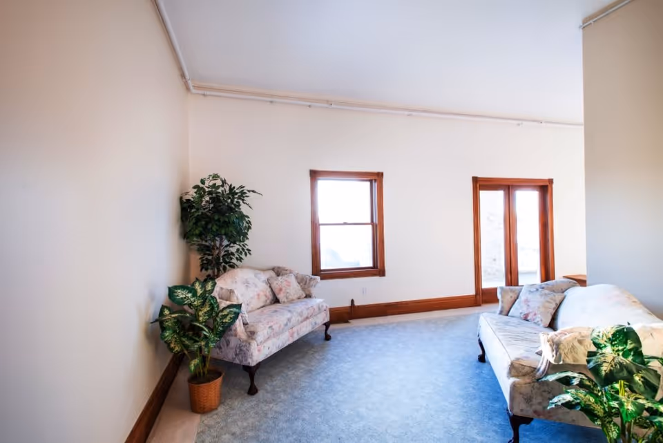 Bright living room with two floral sofas, potted plants, wood-trimmed windows and blue carpeting.