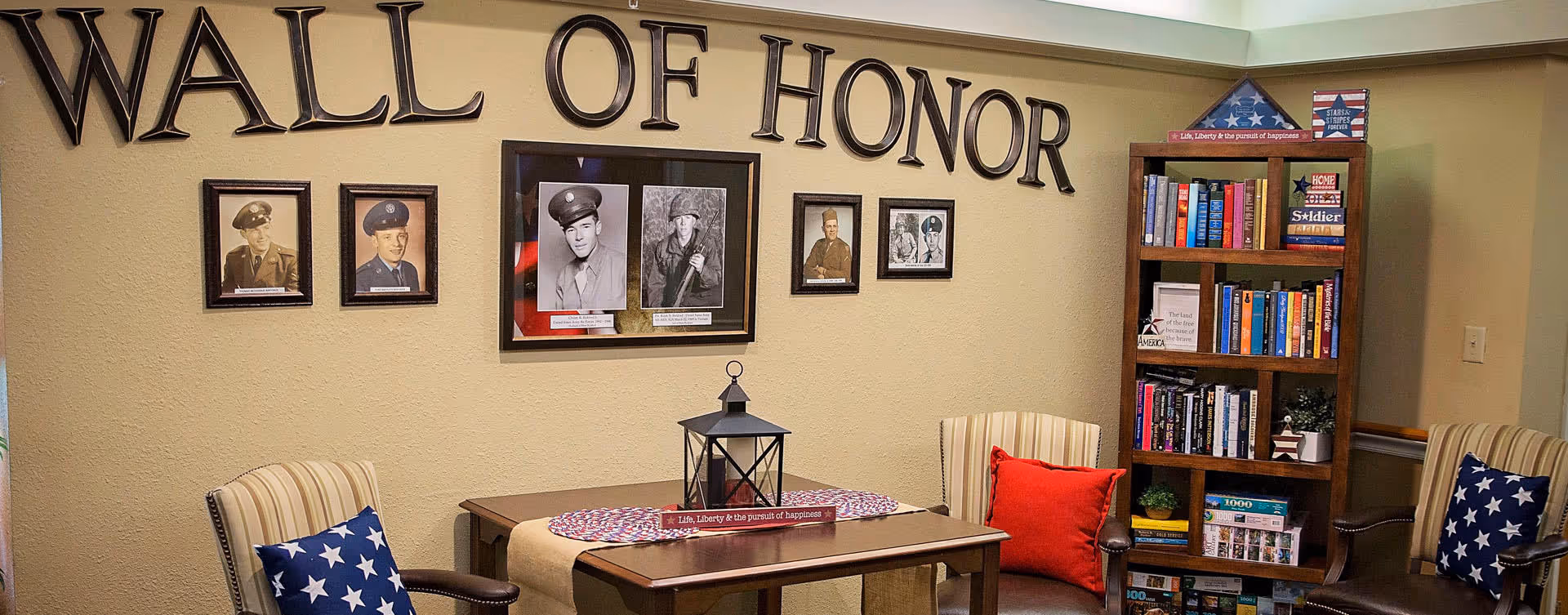 A cozy room with a 'Wall of Honor' display featuring framed black and white photos of military personnel on a beige wall. Below the display is a wooden table with a lantern and a decorative runner. Two armchairs with patriotic star-patterned and red pillows flank the table. To the right, a wooden bookshelf filled with books and patriotic decor items is visible.