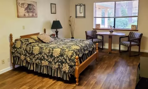 A furnished bedroom with a wooden bed covered by a floral-patterned bedspread, a nightstand and a small table with two chairs by a window.