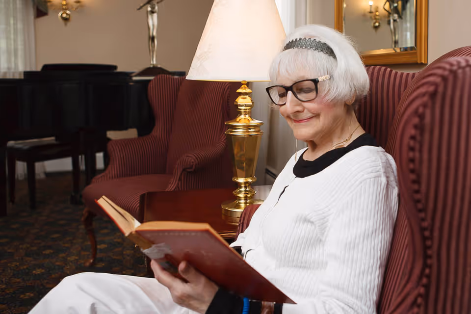 An elderly woman with white hair and glasses is sitting comfortably in a red striped armchair, reading a book. A brass table lamp with a white shade is on a wooden side table next to her. In the background, there is another matching armchair and a black grand piano.